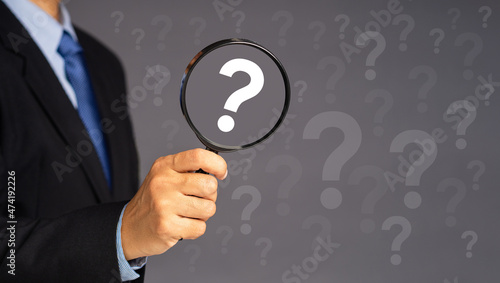 A businessman in a suit holding a magnifying glass look at a white question mark symbol while standing over a gray background. Business, research, and question mark concept
