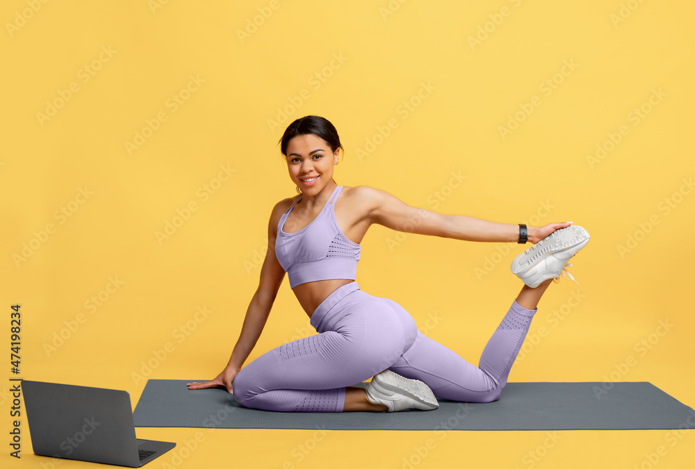 Fototapeta premium Training on isolation. Happy african american lady exercising and watching online tutorials on laptop, yellow background