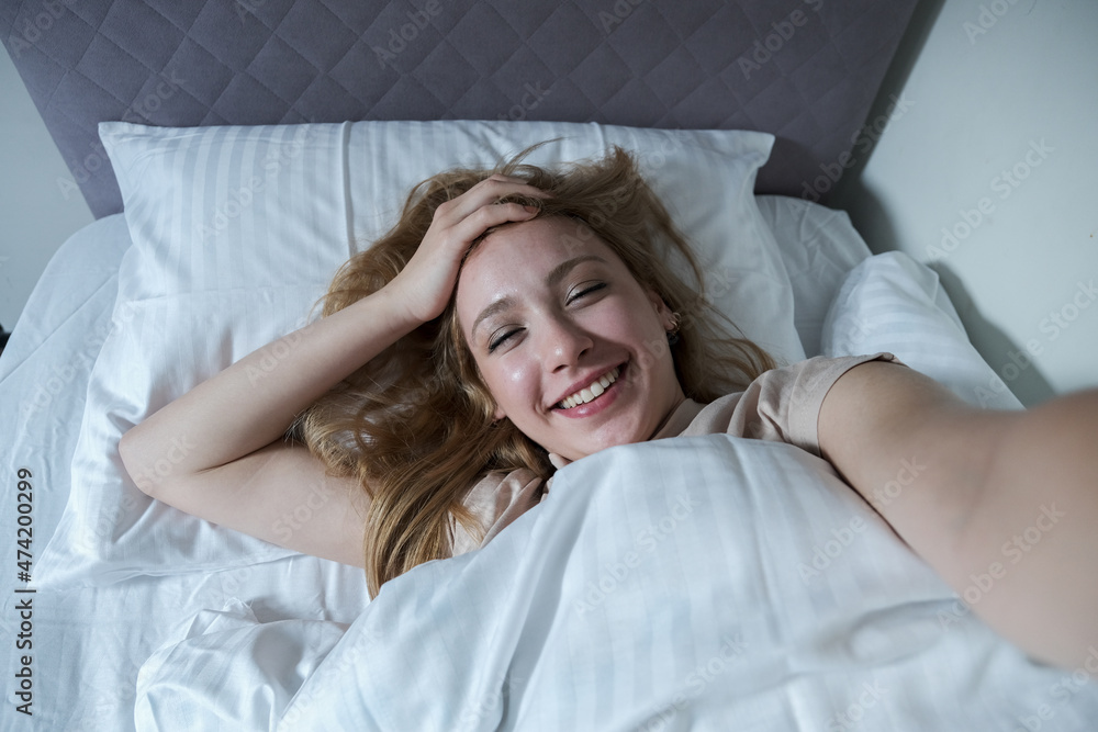 Young blond woman taking a smiling selfie on her bed with a white cover, newly awakened woman taking selfie Young blond woman taking a smiling selfie on her bed with a white cover, newly awakened woman taking selfie
