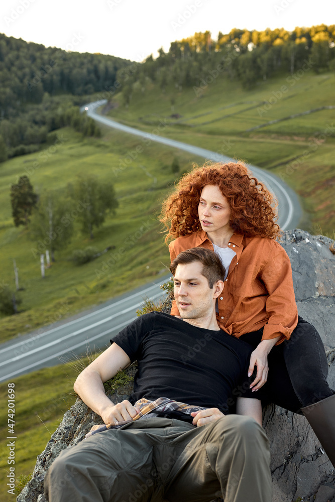 Relaxed Caucasian Couple Sitting On The Head Of Mountain, Having Rest After Climbing, Looking At Side In Contemplation of Nature Landscape Around. Road In The Background. Man And Redhead Woman