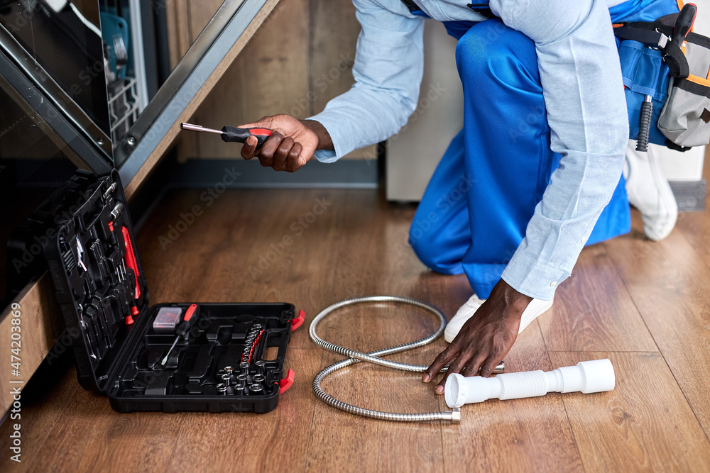 Cropped Professional Handyman Of African American Appearance In Blue Overalls Repairing Dishwasher, Using Instruments Opening Toolbox, In Kitchen Floor At Home. Close-up hands