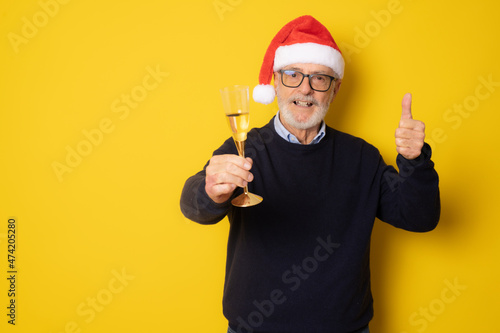 Senior man in santa hat drinking champagne with thumb up standing isolated over yellow background. © Danko