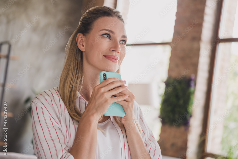 Photo of dreamy young blond lady hold telephone look wear striped shirt at home in living room alone Photo of dreamy young blond lady hold telephone look wear striped shirt at home in living room alone