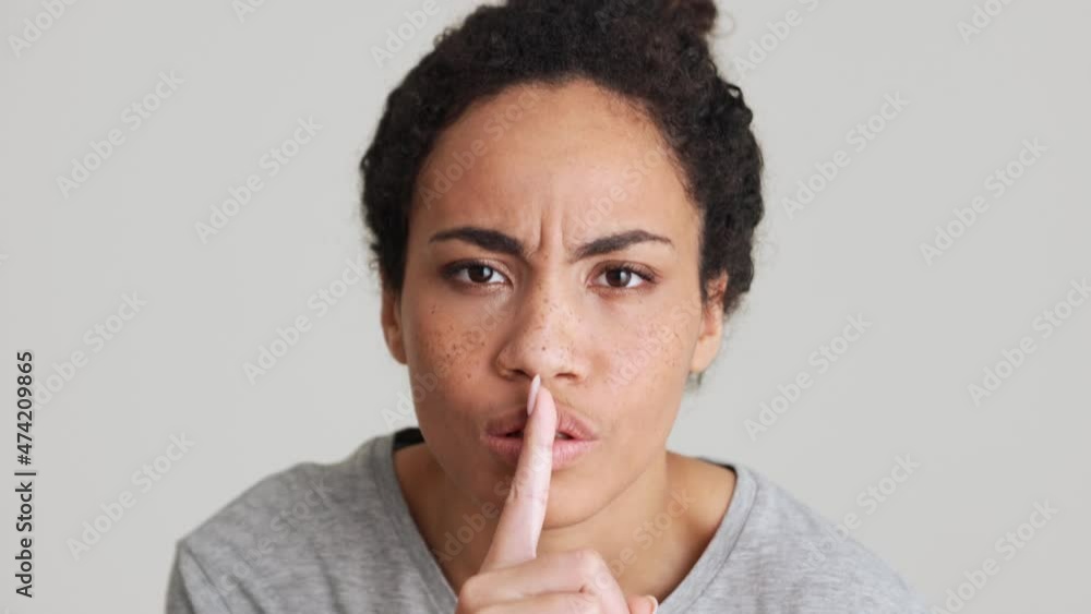 Displeased African woman in t-shirt showing silence gesture in the grey studio Displeased African woman in t-shirt showing silence gesture in the grey studio