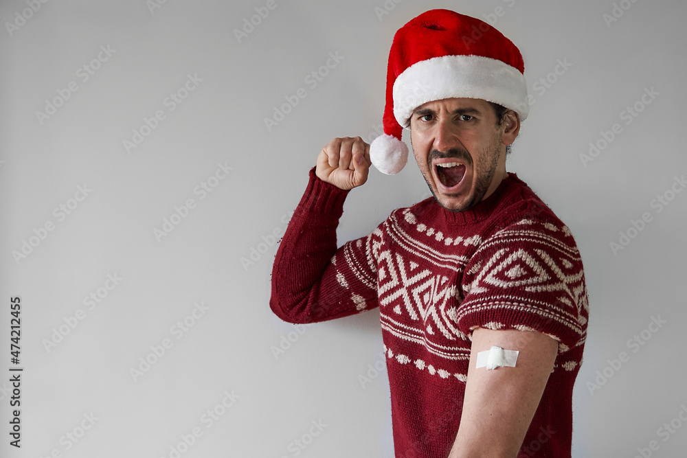 vaccine strong for Christmas. Hyped young man poses with santa claus hat and red sweater with arm bandage over silver grey background vaccine strong for Christmas. Hyped young man poses with santa claus hat and red sweater with arm bandage over silver grey background