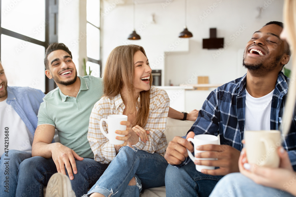 Group of happy multiracial friends drinking coffee in living room, having conversation, enjoying time together