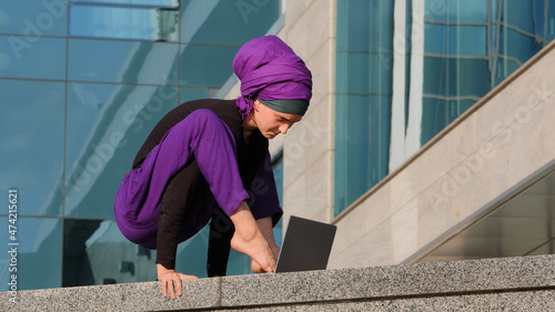 Young strong business woman islamic muslim student stands in balance handstand asana acrobatic movement typing with feet on laptop city building background closes computer finishing work resting
