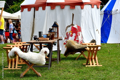 A close up on the display of various medieval items of eqipment, including tables, chairs, lamps, and shields located in the middle of the field next to a colorful cloth tent seen in Poland in summer
