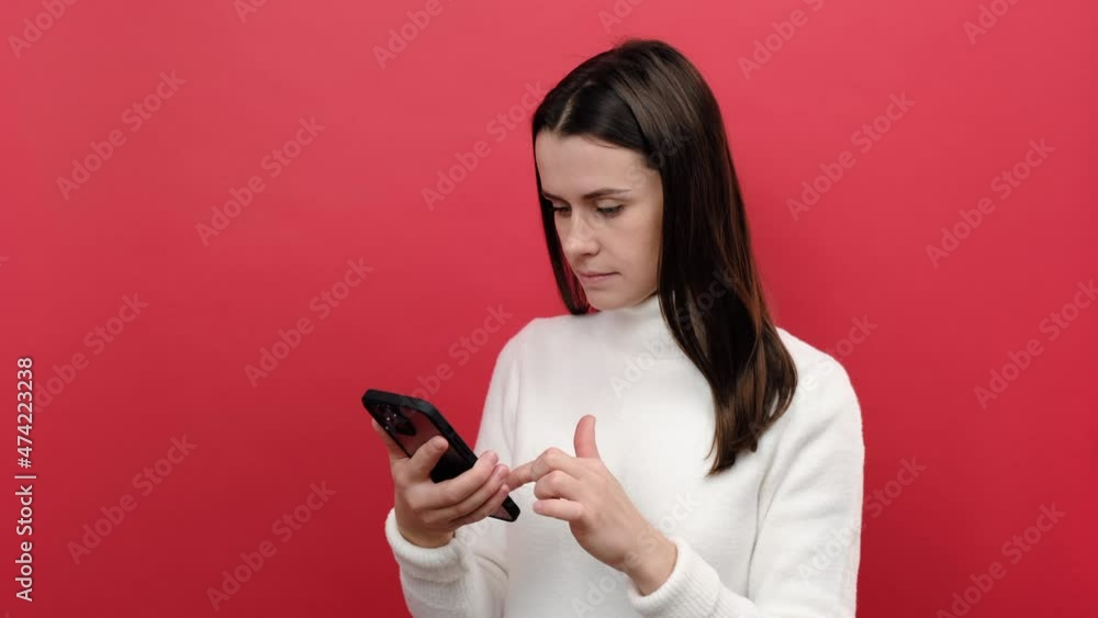 Portrait of cute young brunette woman using cell phone, smiling while scrolling web, satisfied with mobile application, wears cozy white sweater, posing isolated over red color background in studio Portrait of cute young brunette woman using cell phone, smiling while scrolling web, satisfied with mobile application, wears cozy white sweater, posing isolated over red color background in studio