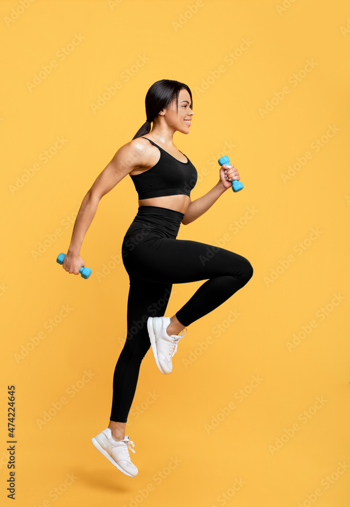 Workout concept. Full length shot of young black woman in sportswear exercising with two dumbbells, yellow background