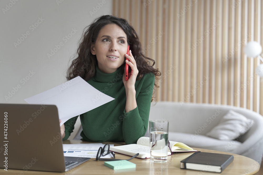 Smiling Italian businesswoman sitting at table making call at her workplace while working on laptop Smiling Italian businesswoman sitting at table making call at her workplace while working on laptop