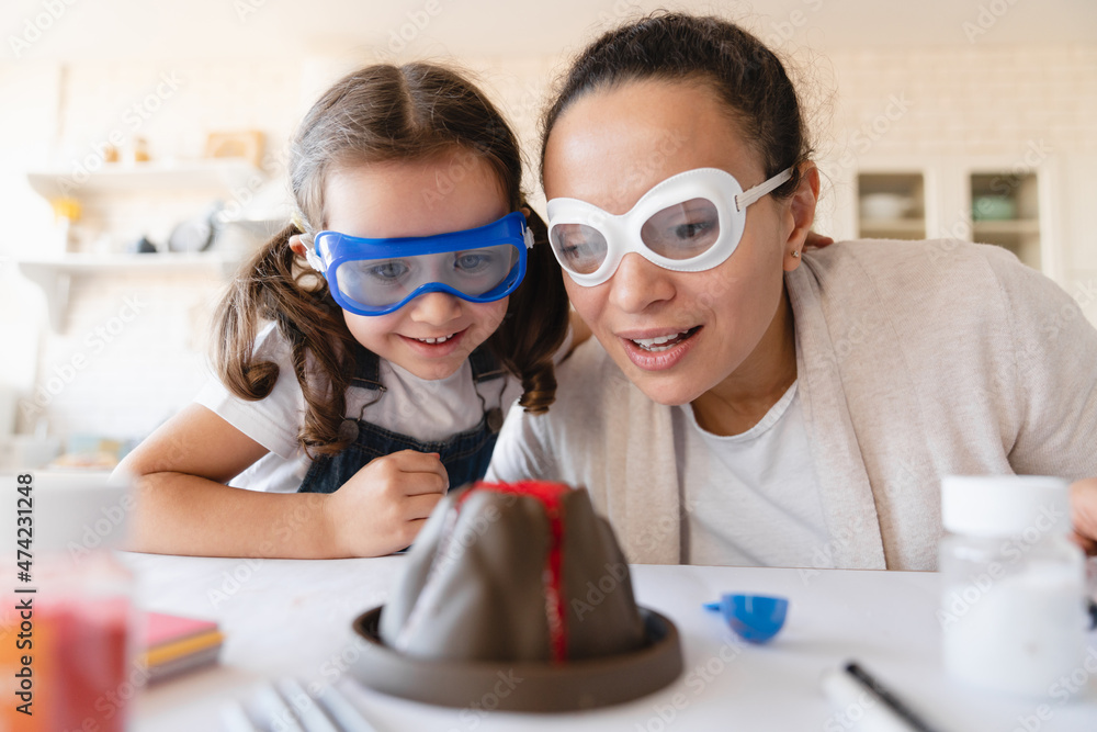 Closeup cropped shot of mother and daughter in protective eyewear watching volcano baked soda eruption at home kitchen together, making school science project. Home chemistry lab - obrazy, fototapety, plakaty 