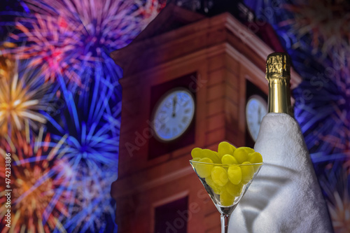 Glass with grapes on the background of a blurry bottle of champagne, clock tower in twelve o'clock and fireworks. Spanish tradition of eating twelve grapes to celebrate New Year.