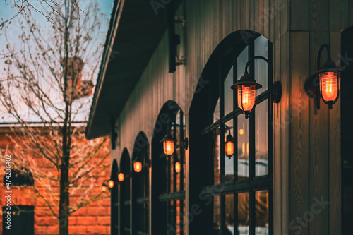 One-story building. Row of black arched windows in dark wooden wall. Vintage lanterns close-up. Orange stone house and leafless bare tree branches covered by snow at background.