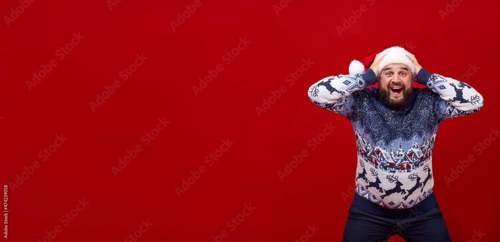 Banner. The man is holding on to the New Year's hat. Emotional bearded man on a red background. Sweater with deer on the guy. Banner. The man is holding on to the New Year's hat. Emotional bearded man on a red background. Sweater with deer on the guy.