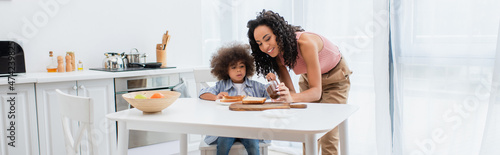 Happy african american woman holding chocolate paste near kid and bread in kitchen, banner.