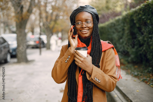 Young black woman in a coat drinking coffee outside and using a phone