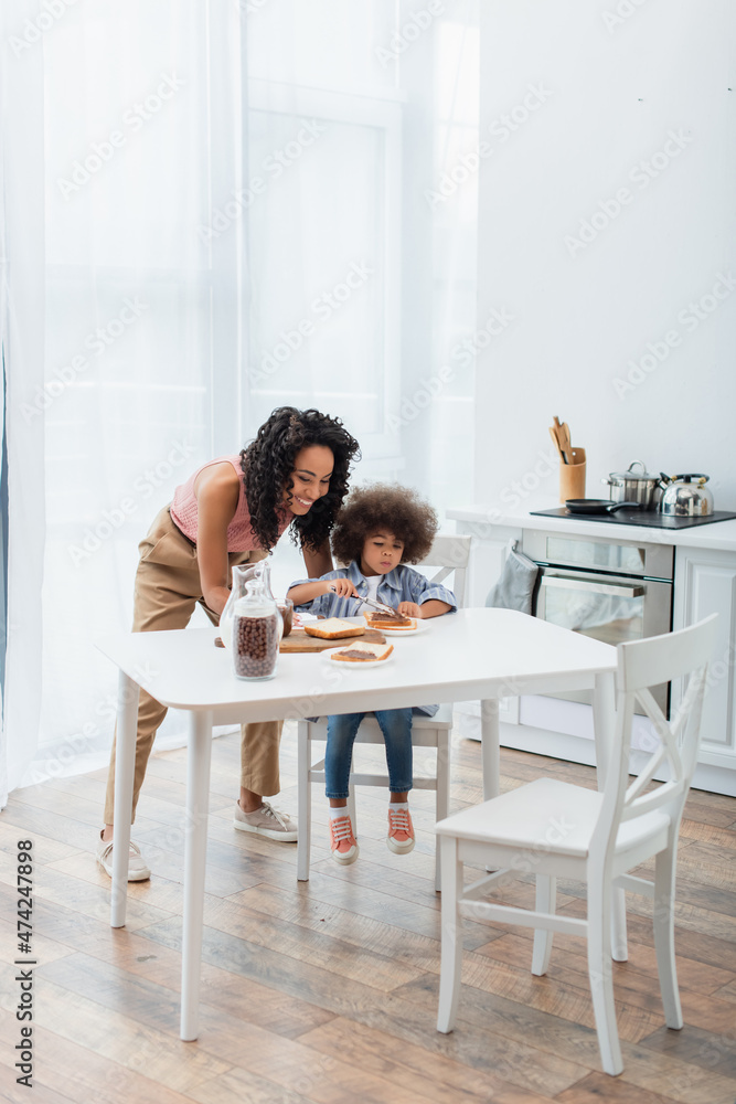 Smiling african american mom looking at kid spreading chocolate paste on bread and cereal at home.