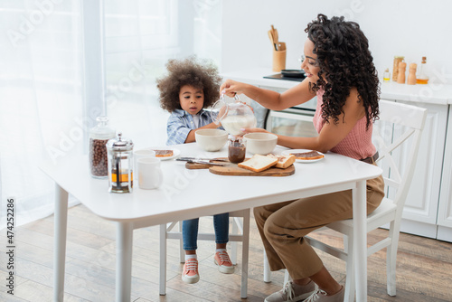 African american mom pouring milk near breakfast and daughter at home.