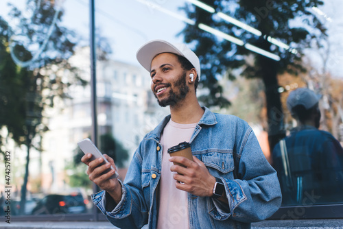 Smiling Spanish hipster guy in bluetooth headphones enjoying positive music playlist from mobile radio using 4g wireless in city, millennial male tourist in earbuds holding coffee to go and smartphone photo