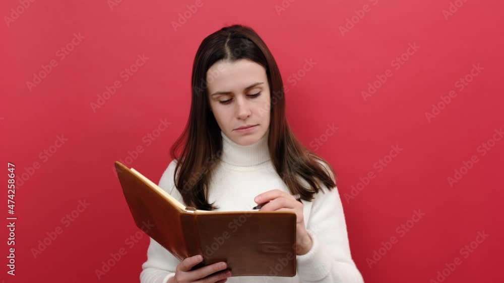 Portrait of pensive young brunette woman 20s making notes, writing down thoughts with pencil into notepad, to do list, wearing white knitted sweater, posing isolated over pastel red background studio Portrait of pensive young brunette woman 20s making notes, writing down thoughts with pencil into notepad, to do list, wearing white knitted sweater, posing isolated over pastel red background studio