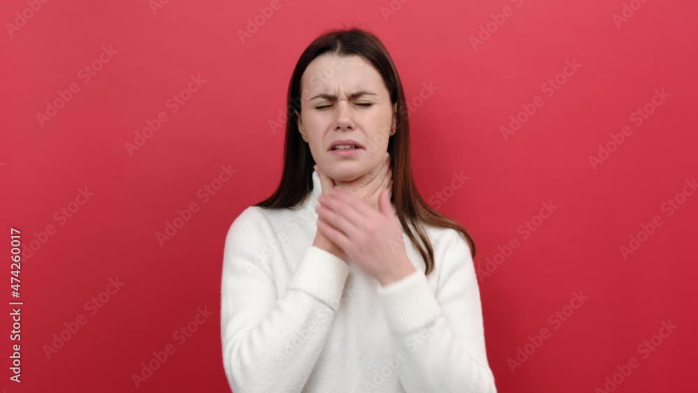 Portrait of unhappy young female 20s wincing in pain, touching painful neck, suffers sore throat, virus or tonsillitis, wears white sweater, posing isolated over red color background wall in studio Portrait of unhappy young female 20s wincing in pain, touching painful neck, suffers sore throat, virus or tonsillitis, wears white sweater, posing isolated over red color background wall in studio