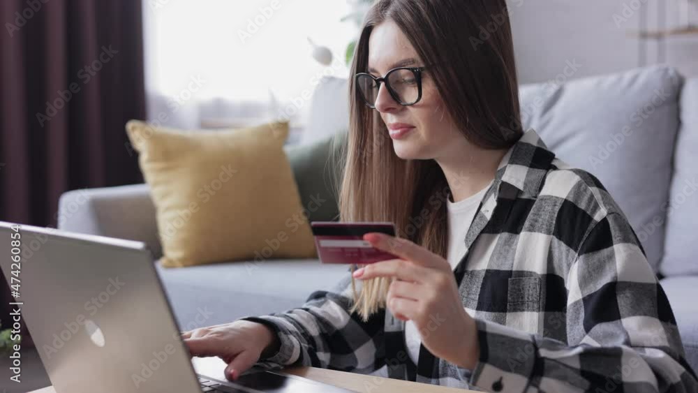Close up of the Caucasian young woman in glasses shopping online on the computer while sitting in living room at home and holding credit card. Finance credit banking money transaction. Close up of the Caucasian young woman in glasses shopping online on the computer while sitting in living room at home and holding credit card. Finance credit banking money transaction.
