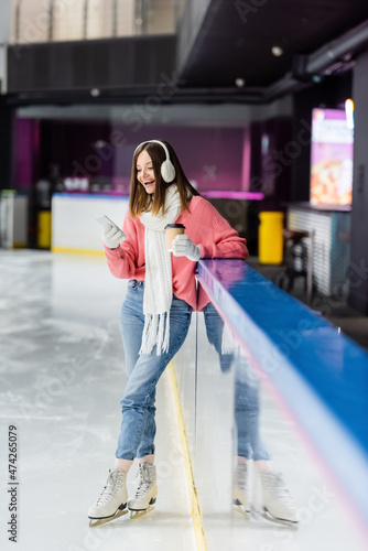 full length of young woman with paper cup and smartphone standing on ice rink. © LIGHTFIELD STUDIOS