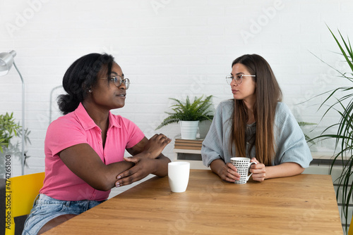 Two best friends hanging out at home together and having fun while sitting in cozy modern apartment, drinking coffee. Two women gossip about the men they met last night out at the party in the club.