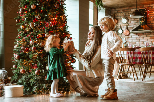 family pregnant mom and two siblings little boy and girl in stylish clothes in chalet are ready to welcome Christmas and New Year near Christmas tree with presents with table set for festive dinner