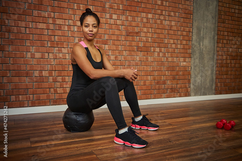 Beautiful African athlete woman in stylish black activewear sitting on a medicine ball and looking at camera at gym studio