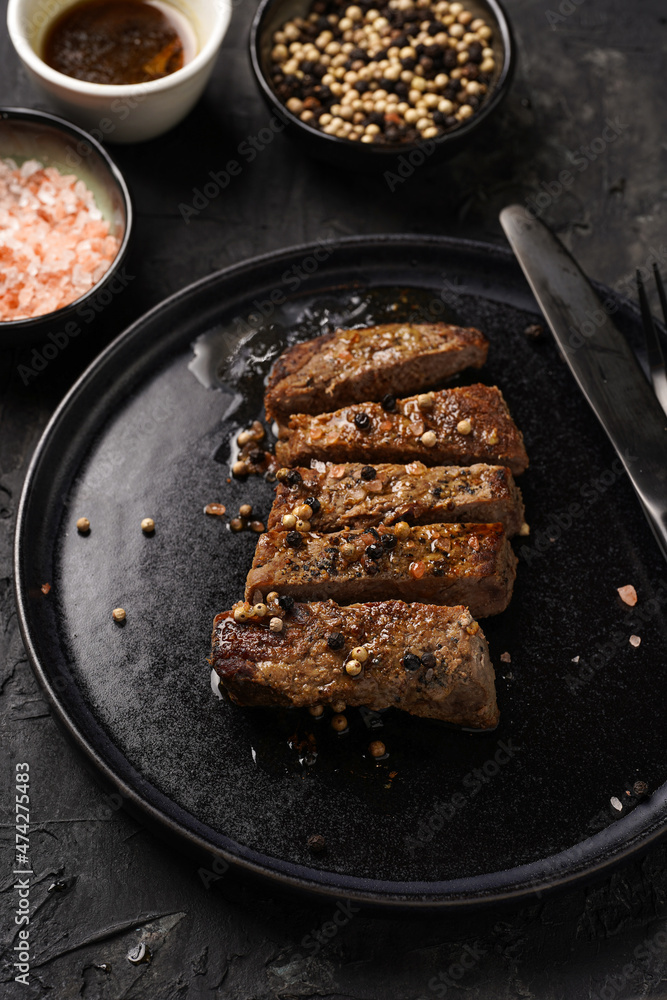 Fototapeta premium A piece of cooked beef meat steak on black plate cut in slices, white and black peer, coarse sea salt