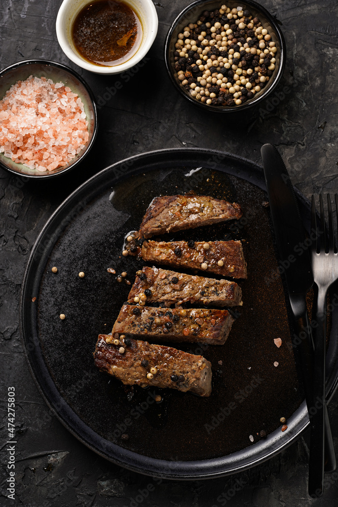 A piece of cooked beef meat steak on black plate cut in slices, white and black peer, coarse sea salt A piece of cooked beef meat steak on black plate cut in slices, white and black peer, coarse sea salt