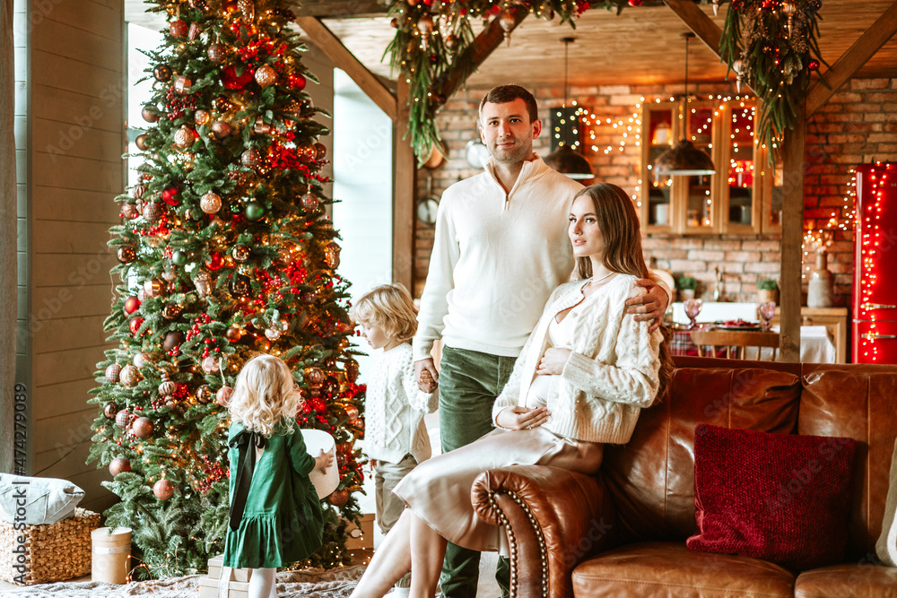family of four parents pregnant mom and dad and two siblings little boy and girl in stylish clothes in chalet are ready to welcome Christmas and New Year near Christmas tree with gift box presents