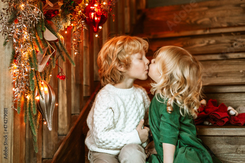 siblings children boy and little girl with blond hair and blue eyes in beautiful clothes sit in living room on stairs near Christmas tree for family celebration of Christmas and New Year, kiss and hug