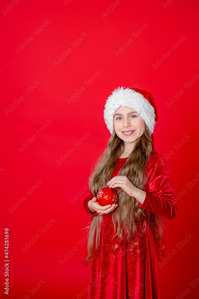 Portrait of a cheerful cheerful girl in a Santa hat with a Christmas ball, isolated on a bright red background. Portrait of a cheerful cheerful girl in a Santa hat with a Christmas ball, isolated on a bright red background.