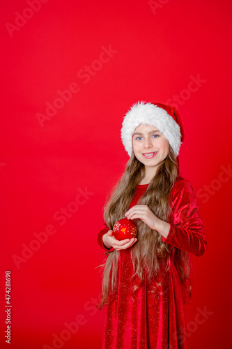 Portrait of a cheerful cheerful girl in a Santa hat with a Christmas ball  isolated on a bright red background. 