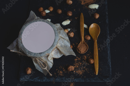 High angle view of hazelnut nut butter jar with empty label design, with spoons full of carob powder and honey on black background