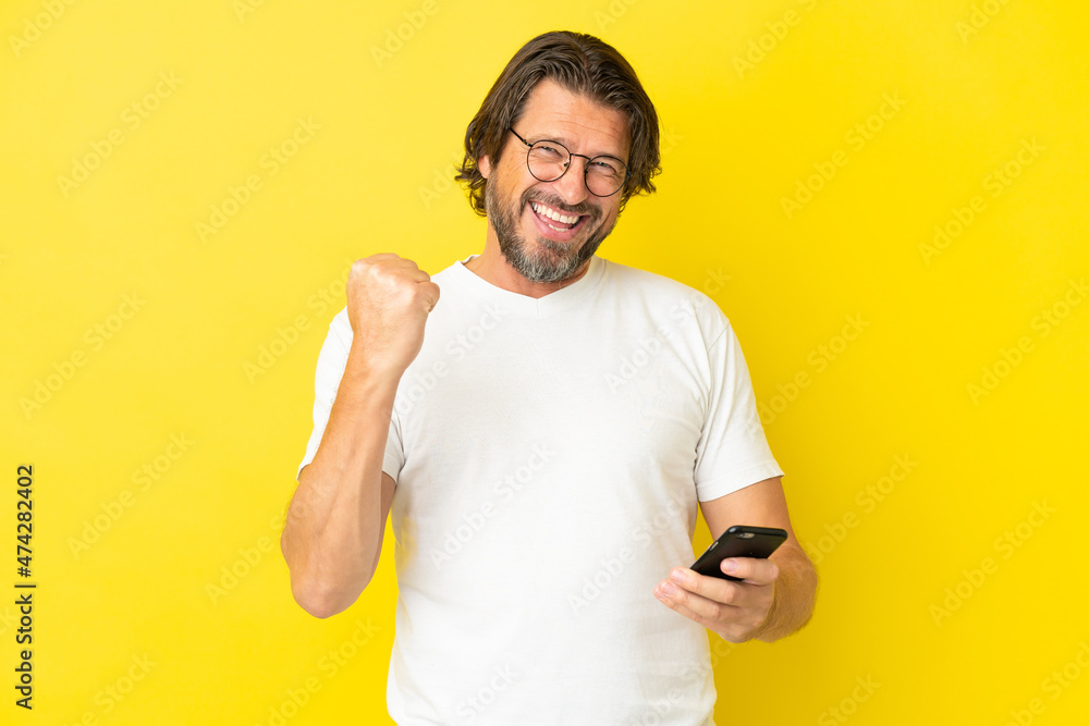 Senior dutch man isolated on yellow background with phone in victory position Senior dutch man isolated on yellow background with phone in victory position