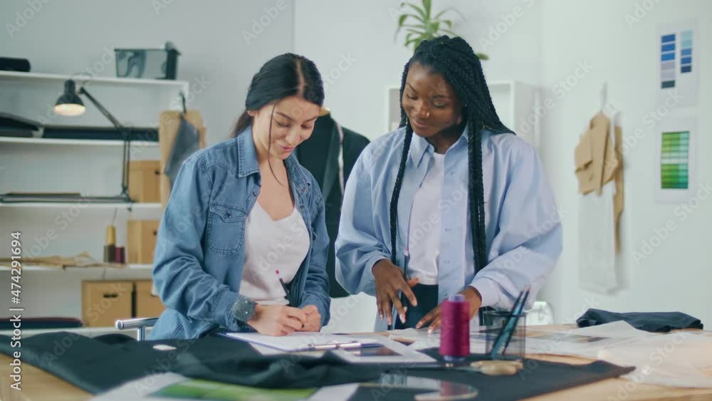 Successful Fashion Designers Caucasian Woman and African-American Woman Create a New Fashion Collection in a Sewing Workshop. Handicraft, Creative Small Business. Apparel Design Startup Concept. Successful Fashion Designers Caucasian Woman and African-American Woman Create a New Fashion Collection in a Sewing Workshop. Handicraft, Creative Small Business. Apparel Design Startup Concept.