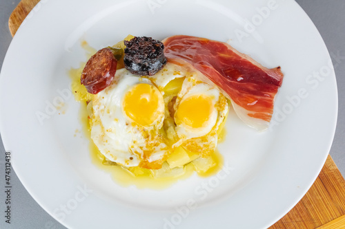Fried eggs with serrano ham, fries, chorizo and blood sausage dish plate. Typical Mediterranean food Spain tapas. Alpujarreño tapa background. Broken fried eggs with potatoes and Iberian cured ham. photo