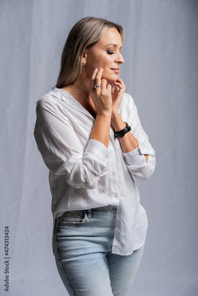 beautiful woman portrait in studio posing standing. girl in a white shirt and blue jeans, barefoot. blonde with straight hair and bright makeup. beauty concept. Gray background beautiful woman portrait in studio posing standing. girl in a white shirt and blue jeans, barefoot. blonde with straight hair and bright makeup. beauty concept. Gray background