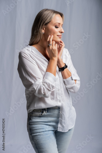 beautiful woman portrait in studio posing standing. girl in a white shirt and blue jeans, barefoot. blonde with straight hair and bright makeup. beauty concept. Gray background
