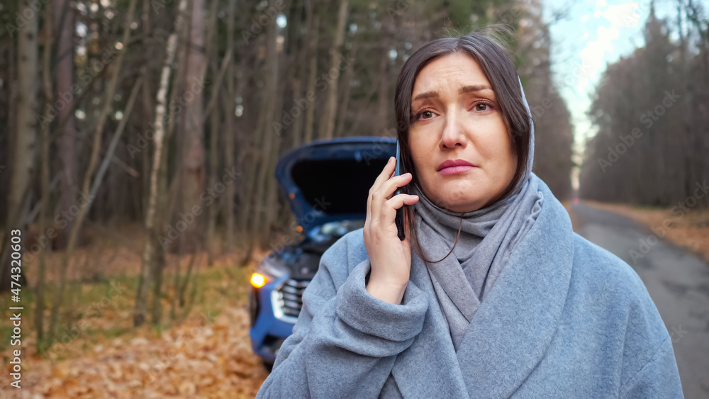 Lady driver in warm coat stands near broken automobile with open hood and calls emergency repair service for help on roadside near autumn forest, closep Lady driver in warm coat stands near broken automobile with open hood and calls emergency repair service for help on roadside near autumn forest, closep