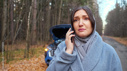 Lady driver in warm coat stands near broken automobile with open hood and calls emergency repair service for help on roadside near autumn forest, closep photo
