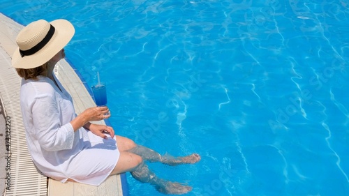 relaxing senior woman dangling her legs in an outdoor swimming pool holding a blue refreshing cocktail wearing a straw hat. Summer rest. All inclusive