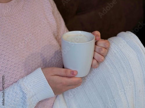 A girl in a knitted light sweater holds a white mug of cocoa in her hands, her legs are covered with a knitted white blanket, she warms up in cold weather with a hot drink