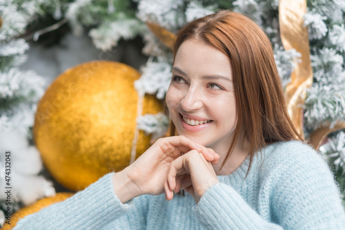 New Year close up portrait of beautiful smiling caucasian young woman in cozy wool warm light blue sweater. Christmas tree on the background.