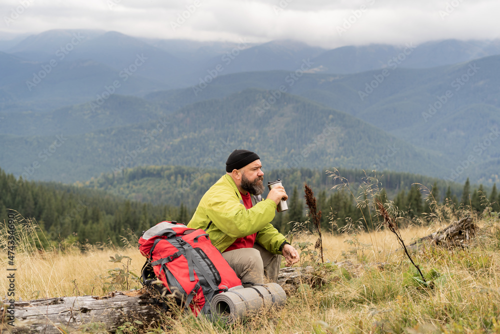 Fototapeta premium a bearded Caucasian male tourist on vacation while hiking in the mountains sits on the trunk of a fallen tree and drinks tea or coffee from a thermos cup. Halt in the mountains.