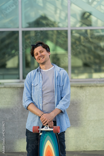 Young skateboarder guy over modern glass wall background, street portrait of hipster skater holding longboard wearing trendy denim shirt and bandana look in camera. Urban youth trend lifestyle concept